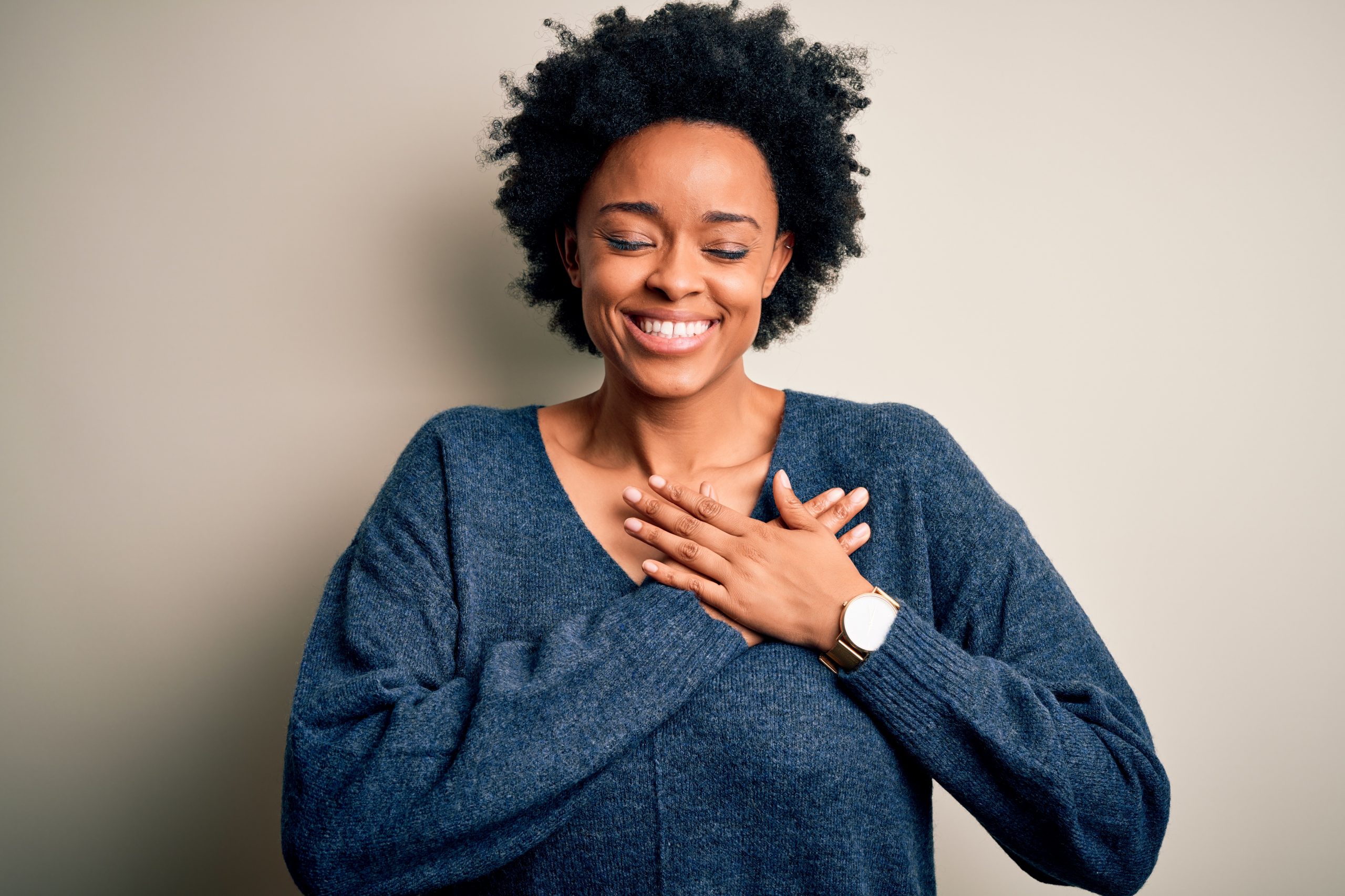 Young beautiful African American afro woman with curly hair wearing casual sweater smiling with hands on chest with closed eyes and grateful gesture on face. Health concept.